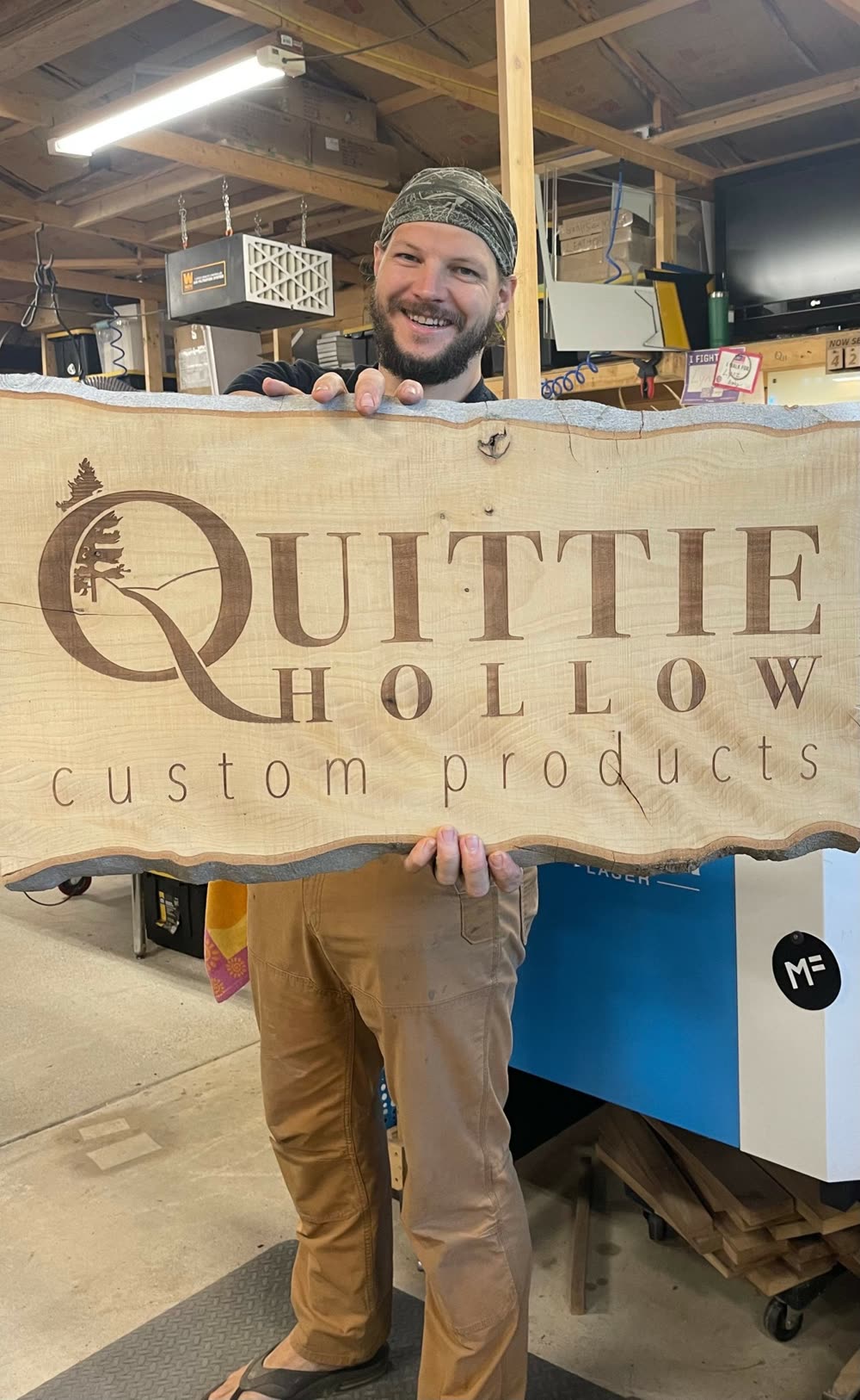 Mike Reese in the Quittie Hollow workshop holding a live-edge slab engraved with the workshop logo, a laser cutter behind him